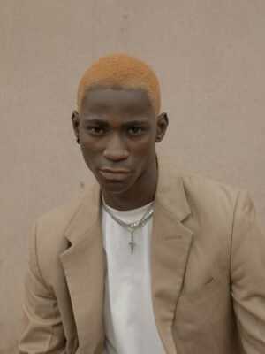 Confident African American male with dyed hair wearing trendy suit and chain looking at camera while standing near wall on street