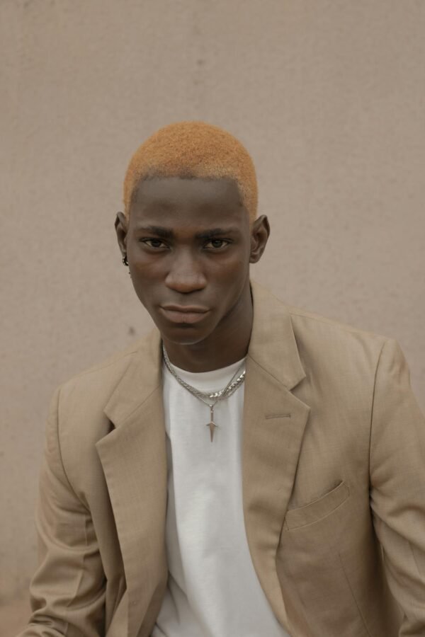 Confident African American male with dyed hair wearing trendy suit and chain looking at camera while standing near wall on street