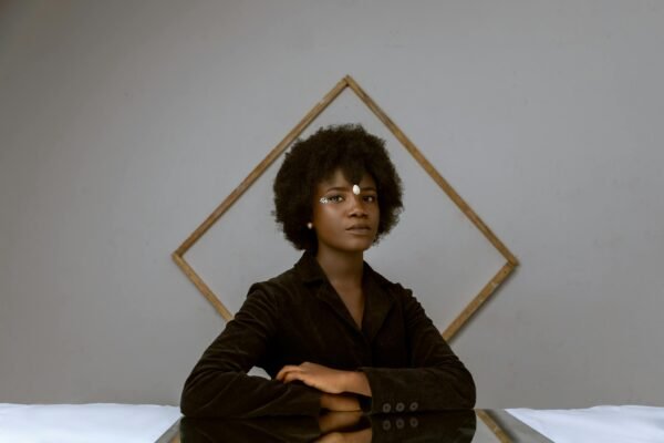 Serious young African American female in classy jacket with creative hairstyle sitting near bed against wall with geometric decor